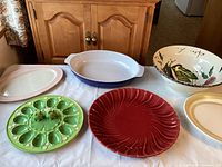 Overview of all ceramic serving dishes displayed on cloth, showing frog egg plate, Emile Henry baking dish, painted vegetable bowl, and white plates.