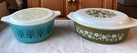 Two vintage Pyrex casserole dishes with lids, turquoise and olive green, shown side by side to display color and lid pattern differences.