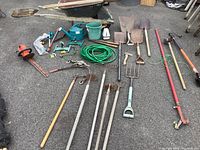 Full view of assorted yard and gardening tools laid out on pavement, including hand tools, rakes, shovels, hose, and wheelbarrow.