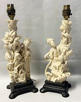 Front view of two ivory colored carved resin lamps with Chinese figures and floral motifs on black bases. Lamp fittings on top visible.