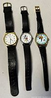 Three vintage watches on a white surface, including two Disney character watches and one traditional watch with yellow case and black strap