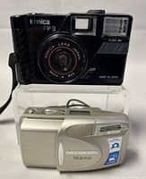 Front view of Konica FP3 black 35mm film camera with lens, flash, ISO markings, and Olympus Stylus 80 silver compact camera stacked below.