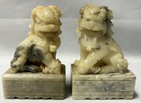 Front view of the pair of vintage carved stone Foo Dog sculptures on bases, showing coloration and wear.