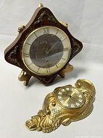 Diamond-shaped vintage KAISER wooden clock with brass corner details and circular glass face showing Roman numerals on a white dial, accompanied by smaller ornate gold-colored PREXIM clock with sculpted leaf and scroll design.
