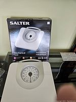 Mechanical bathroom scale in white with a large circular dial and black/red markings, shown in front of its original Salter branded box.