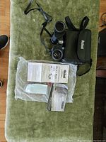 Bushnell Perma-Focus binoculars, carrying case, instruction manual, and lens cloth displayed on green surface.