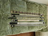 Seven metal and leather ladies belts laid out on a green cloth showing various buckle and belt designs.