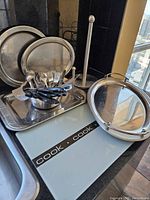 Multiple stainless steel trays, a colander holding black-handled flatware, a glass cutting board with cook branding, and a tall stainless steel paper holder displayed on a countertop near a window.