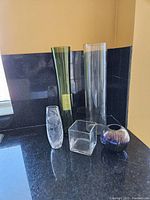 Five assorted decorative glass vases including tall green and clear cylindrical vases, small clear oval vase, clear cube vase, and short round iridescent vase on a countertop.