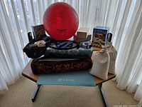 Full view of all items including two yoga mats, Thera-Band ball, wooden blocks, Pilates ball box, resistance bands, weights, hand roller, and cloth bags on table in front of sheer curtains.