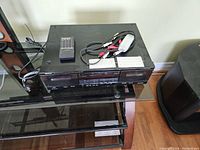 Full view of Denon stereo double cassette deck with remote and cables placed on a cabinet.