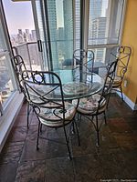 Photo of round glass tabletop with black rod iron base and 5 matching rod iron chairs with floral cushions arranged around it in a bright room with large windows.