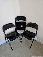 Three black folding metal chairs, two standing open and one folded upright against the wall on carpeted floor.