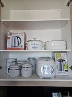 Photo of kitchen shelf with electric kettle in box, canister set, Corning Ware casseroles, ceramic baking dish, rice cooker and coffee butler in box.