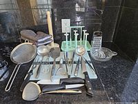 Photo shows mixed kitchen utensils arranged on a countertop including metal spoons, spatulas, strainers, wooden utensils, oven mitts and a paper towel holder.