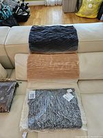 Three Olsen brand shawls displayed on a couch: two textured shawls in dark gray and beige, one shawl still in clear packaging showing a black and white design.