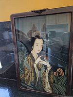 Close up of Oriental woman painting on glass, showing details of clothing, bird and floral background in wood frame