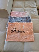 Three pashmina shawls in original packaging laid on a beige couch - peach, pink, and lavender colors visible