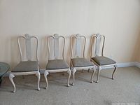 Four white wooden dining chairs with upholstered seats arranged side by side against a wall, carpeted floor.