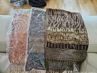 Three faux Pashmina style shawls spread out on a floor or bed. Two are sealed in clear packaging, one is out and fully visible showing brown and tan patterns with fringe.