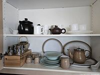 Shelves with a variety of teapots, cups, saucers, mugs, and plates including a dark brown carved oriental-style teapot and multiple white porcelain cups and bowls.