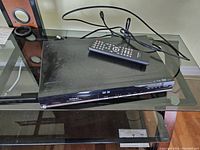 Front and top view of Toshiba DVD and VHS combo player with remote control on top, showing device details and cables behind.