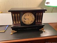 Front view of antique William L. Gilbert parlour clock showing wooden pillars with brass accents, black lacquered wood body, marbleized red molding, and brass feet on a desktop.