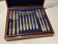 Open wooden case with fitted blue velvet tray holding six ornate fish knives and six matching forks