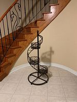 Full view of black metal 3 tier spiral plant stand positioned near wooden stairs, highlighting its height and spiral tier arrangement.