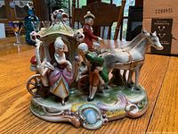 Porcelain figurine showing rococo carriage with two horses and three figures: coachman, gentleman, and lady. Painted in pastel colors with gold detailing on a decorated base.