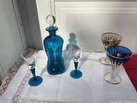 Photo showing decanter with original stopper and two small teal-stemmed liqueur glasses alongside two larger glasses with gold detailing on white cloth background.