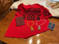 Full view of the red T-shirt with printed Indigenous design, two beaded necklaces, and the maroon-covered Cree language dictionary book laid on a wooden table.