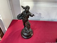 Full front view of metal sculpture of a boy standing with a grape basket on his shoulder and stick over the other shoulder against a red background.