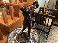Full view of the vintage bronzed metal golfer figurine in a swinging position on a tiled floor surface with wooden stairs in the background.