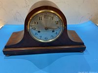 Full view of Tambar German mantle clock wooden case with curved top and wide base on blue background.