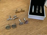 Overview of four pairs of cuff links and one pair of rectangular tie clips on a wooden surface, cuff links include varied shapes and finishes