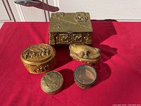 Five brass trinket boxes displayed on red cloth under sunlight, showing shapes and relative sizes.