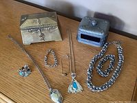 Photo showing brass Benedict Copper jewelry coffin, metal blue glass inset jewelry coffin, various vintage necklaces, bracelet, and Scottish brooch with gemstones.