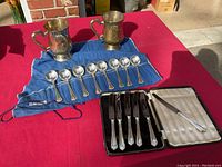 Overview of all items: 2 chased silver plated brass mugs, 8 Wallace cream soup spoons on blue tarnish cloth, and C.P. Walker & Co. Sheffield knives set in case.