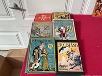 All six Oz books arranged on a red tabletop against a white door background: Wizard of Oz, The Scarecrow of Oz, Kabumpo in Oz, The Lost Princess of Oz, The Lost King of Oz, and Sky Island.