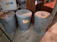 Photo showing three vintage galvanized metal garbage bins with lids and side handles, one bin has a vintage label.