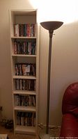 White 6 shelf unit filled with DVDs/CDs next to the torchiere floor lamp in a corner of a room.