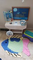 Full view of the lot showing the small white wooden table with items on top and wall behind it, kids stool on right side, and colorful fish rug on the floor