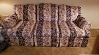 Front view of a large sofa with floral-striped fabric upholstery in cream, mauve, and dark hues, showing cushioned back and seat, and padded arms.