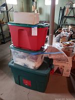Stacked three Christmas storage totes, two with green lids and one with red lid, containing various unidentifiable Christmas decorations, plus a cardboard box on top.