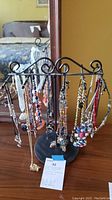 Full view of a black metal ornamental necklace stand holding multiple necklaces in assorted colors and styles, placed on a wooden floor in front of a mirror.
