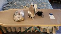 Ironing board with beige cover, iron, black cord and bag of buttons on top, situated on a bed.