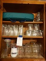 Glassware set including tall and short glasses stacked on wooden shelves, some plastic cups including a Costco-branded tumbler visible, and green carrying case on the top shelf