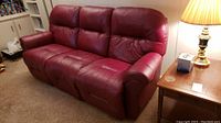 Photo of three-seat deep scarlet leather sofa showing front and side profile with armrest and cushioned backrest, part of living room setting.