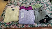 Seven women's sweaters folded on floral bedspread showing a variety of colors and patterns including a grey dog design sweater.